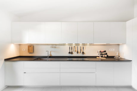 Modern, minimalist kitchen with sleek white cabinets, dark countertops, and hanging utensils.