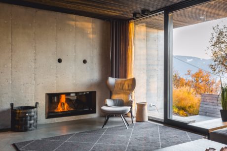 Cozy concrete living space with fireplace, armchair, and panoramic window overlooking autumn landscape.