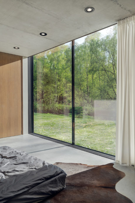 Modern bedroom with large floor-to-ceiling windows offering scenic forest views, concrete ceiling, and minimalist decor.