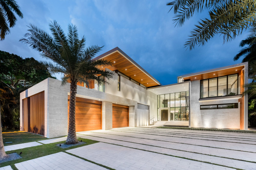 Modern, minimalist exterior with concrete, wood, and glass elements; palm trees frame the entrance.
