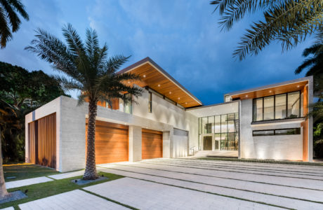 Modern, minimalist exterior with concrete, wood, and glass elements; palm trees frame the entrance.