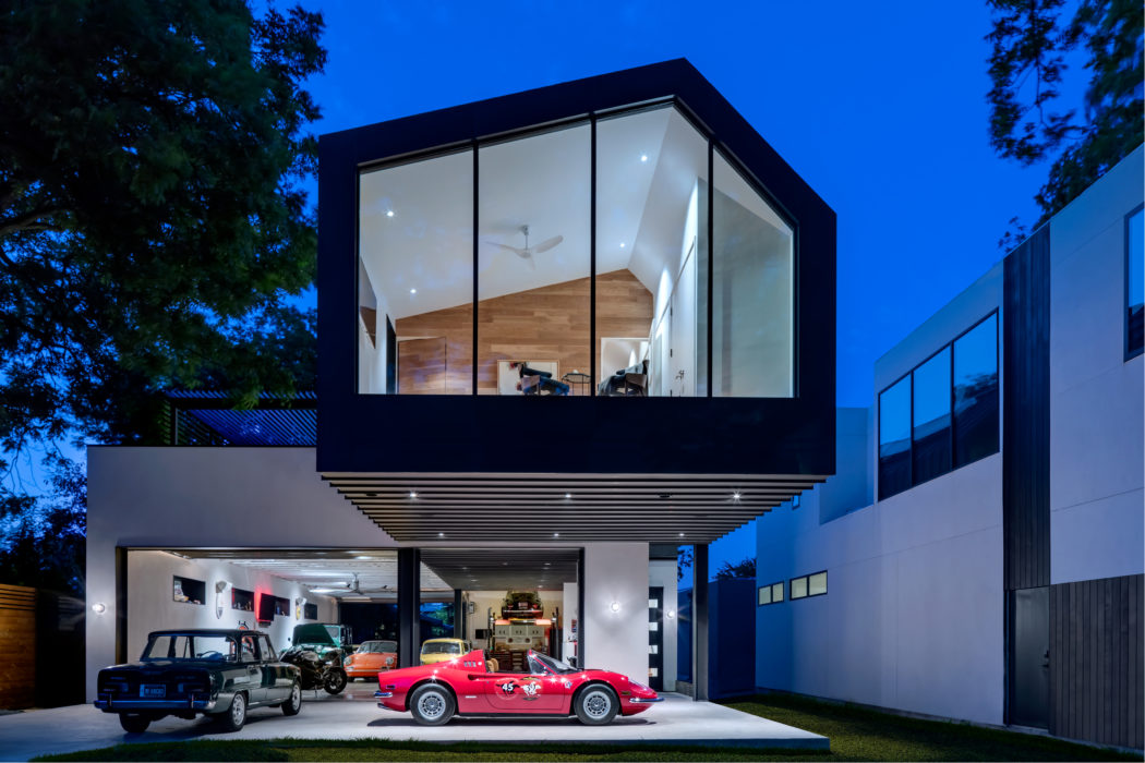 A modern, glass-walled structure with a raised central volume and parked vintage cars below.