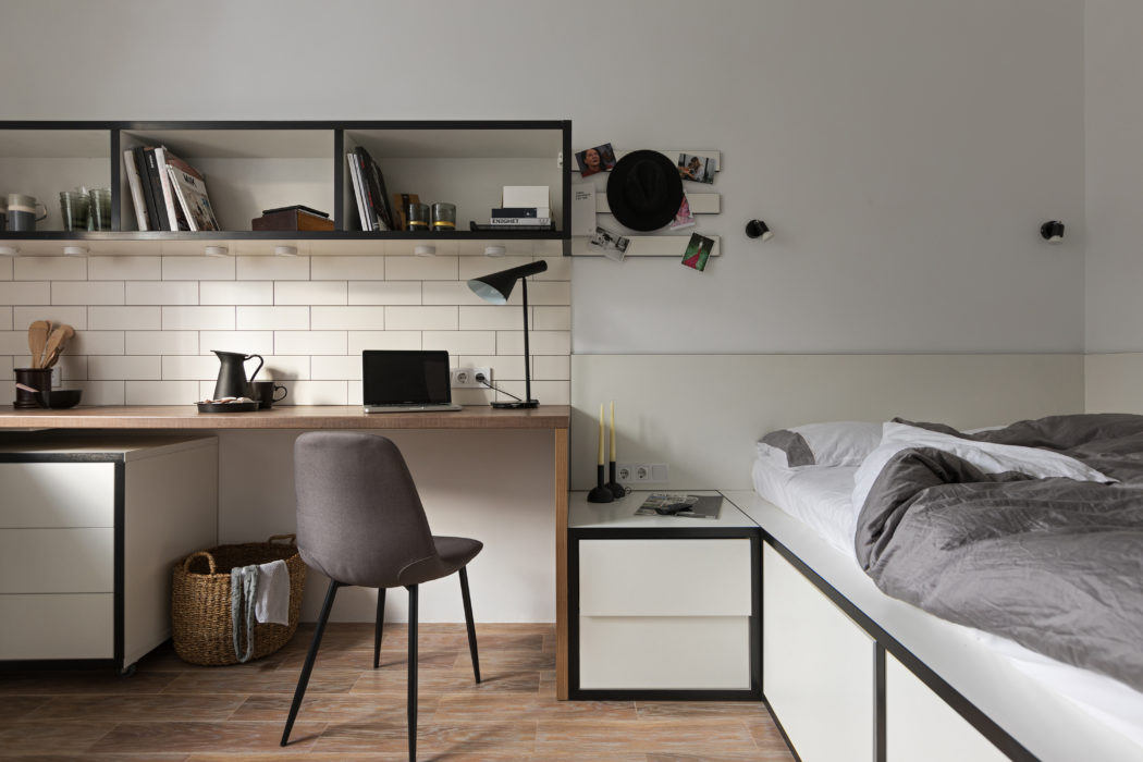 Minimalist studio apartment with wooden desk, shelving, and tiled kitchen area.