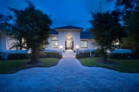Elegant two-story home with arched entrance, brick pavement, and lush landscaping at night.