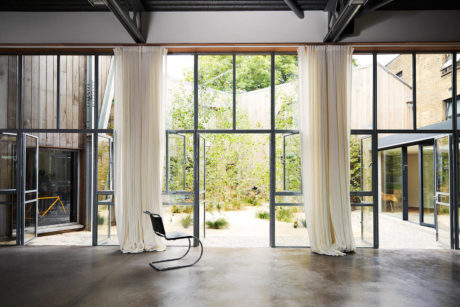 Spacious room with large windows overlooking lush greenery, concrete floor, and minimalist furnishings.