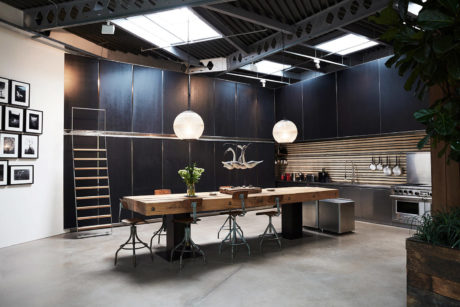 Modern industrial-style kitchen with dark walls, wooden table, and sleek appliances.