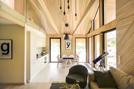 An open-concept, modern cabin-style interior with a vaulted wood-paneled ceiling, minimalist decor, and large windows providing natural light.