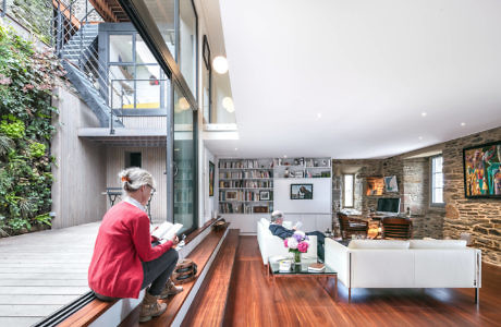 Modern living room with people reading, wooden floors, and large windows.