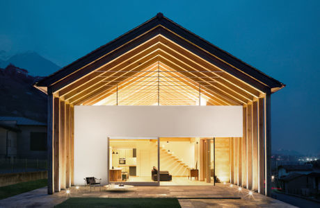 Modern house with illuminated interior and exposed wooden beams at dusk.