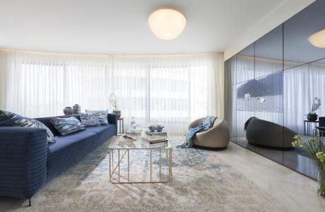 Modern living room with blue sofas, glass table, and sheer curtains.