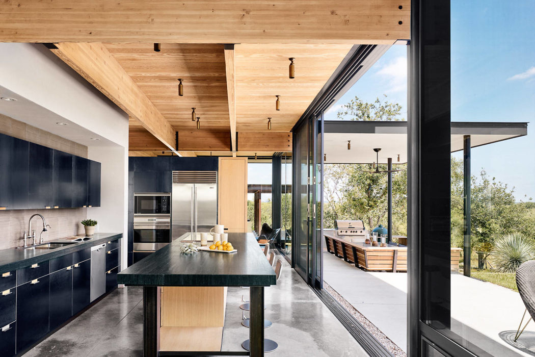 Modern kitchen interior with wooden ceiling and large windows overlooking a patio.