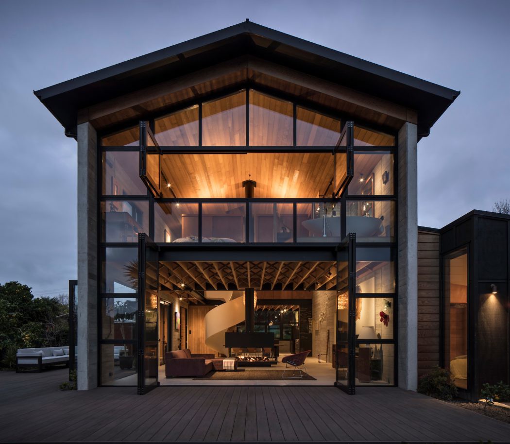 Modern house at dusk with illuminated interior, large glass front, and wooden deck.