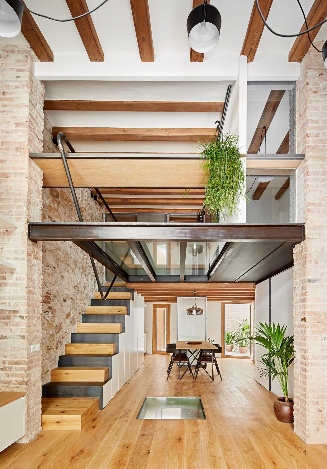 Modern interior with exposed brick, wooden beams, floating staircase, and hanging plants.