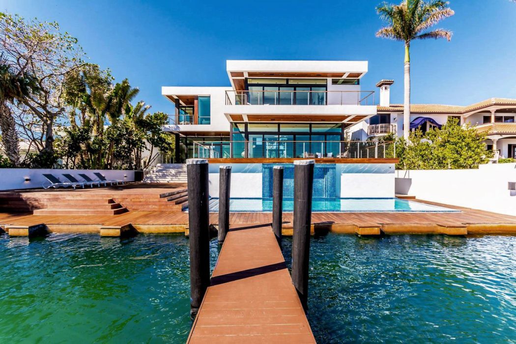 Modern waterfront house with a pier leading to poolside lounging area.