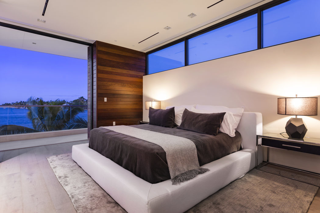 Modern bedroom with large windows overlooking water at dusk.