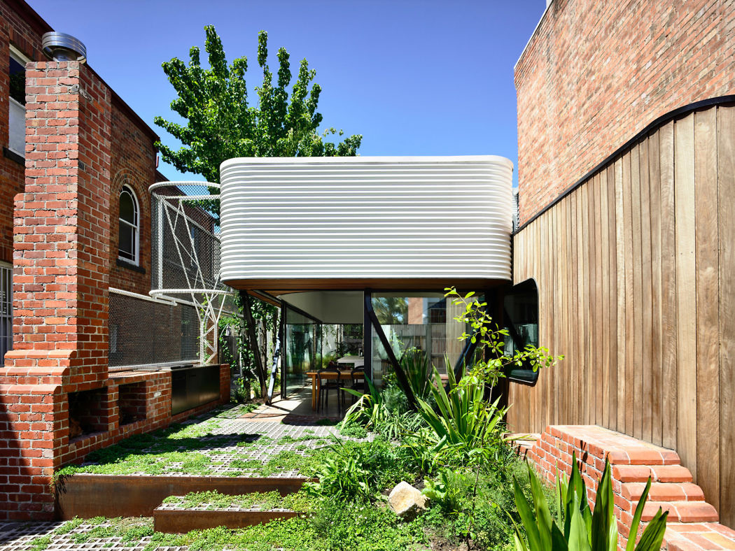 Modern home extension with wavy design, nestled between brick buildings and a wooden fence