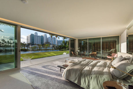 Modern bedroom with large glass windows overlooking a lawn and cityscape.