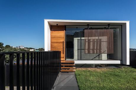 Modern modular home with wooden exterior, glass walls, and a raised porch on a grassy lawn.