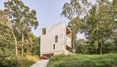 Modern cubic house with white facade amidst green trees with red car parked underneath.