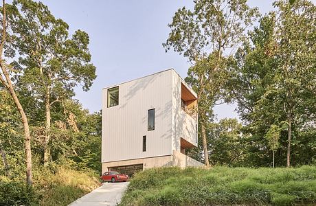 Modern cubic house with white facade amidst green trees with red car parked underneath.