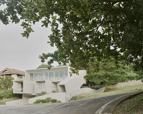 Modern concrete house with large windows, framed by green foliage.