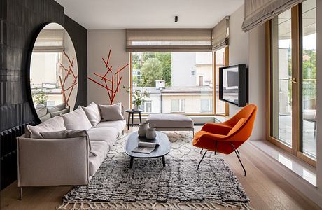 Modern living room with a gray sofa, orange chair, and large windows.