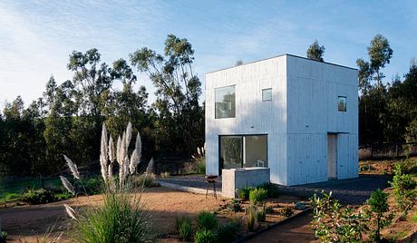 Modern two-story cubic house with large windows, surrounded by natural landscape.