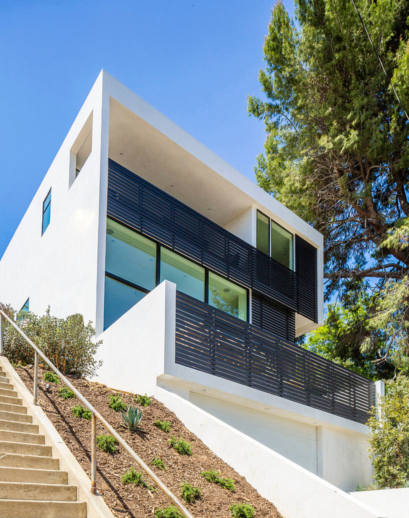 Modern white house with balcony and stairway, surrounded by trees.