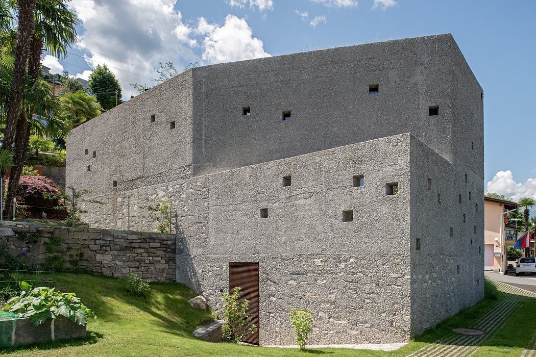 Minimalist concrete building with small windows, set in a green landscape.