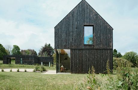 Modern house with a dark wood facade and large window, surrounded by a green lawn