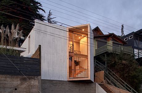 Modern beach house with illuminated interior at dusk, concrete stairs on the side.