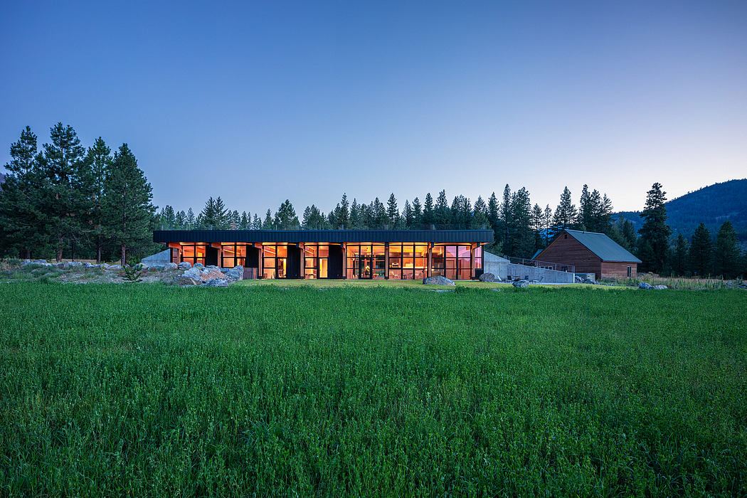 A modern, single-story structure with large windows and warm lighting, surrounded by a grassy field and evergreen trees.