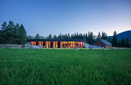 A modern, single-story structure with large windows and warm lighting, surrounded by a grassy field and evergreen trees.