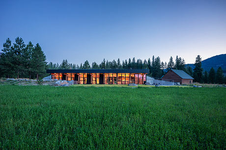 A modern, single-story structure with large windows and warm lighting, surrounded by a grassy field and evergreen trees.