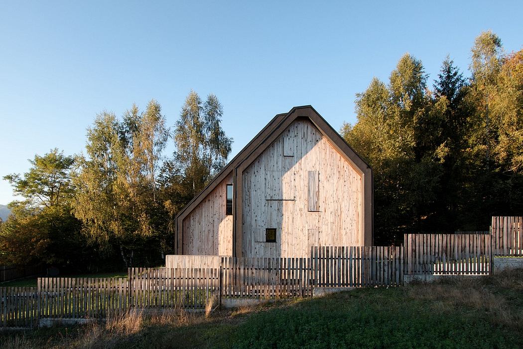 A rustic wooden barn with a pitched roof nestled in an autumnal landscape, surrounded by a wooden fence.