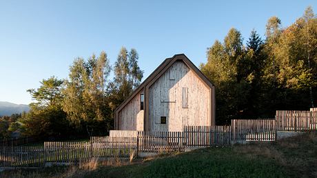 A rustic wooden barn with a pitched roof nestled in an autumnal landscape, surrounded by a wooden fence.