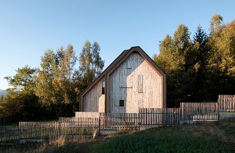 A rustic wooden barn with a pitched roof nestled in an autumnal landscape, surrounded by a wooden fence.