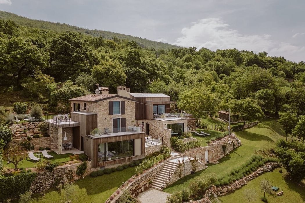 A modern, multi-level stone and wooden structure nestled in a lush, hilly forest landscape.