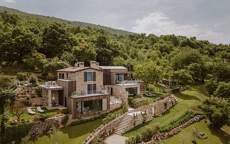 A modern, multi-level stone and wooden structure nestled in a lush, hilly forest landscape.