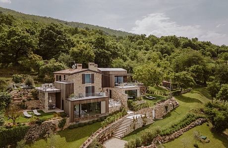 A modern, multi-level stone and wooden structure nestled in a lush, hilly forest landscape.