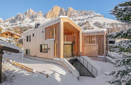 A modern wooden chalet surrounded by snowy mountains and pine trees, with a sleek architectural design.