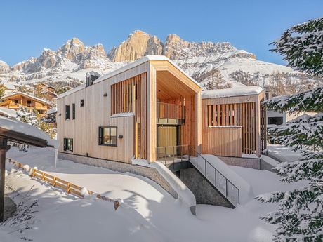 A modern wooden chalet surrounded by snowy mountains and pine trees, with a sleek architectural design.