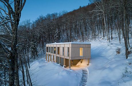 Contemporary cabin with large windows nestled in snowy woods at dusk.