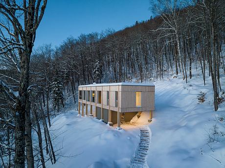 Contemporary cabin with large windows nestled in snowy woods at dusk.