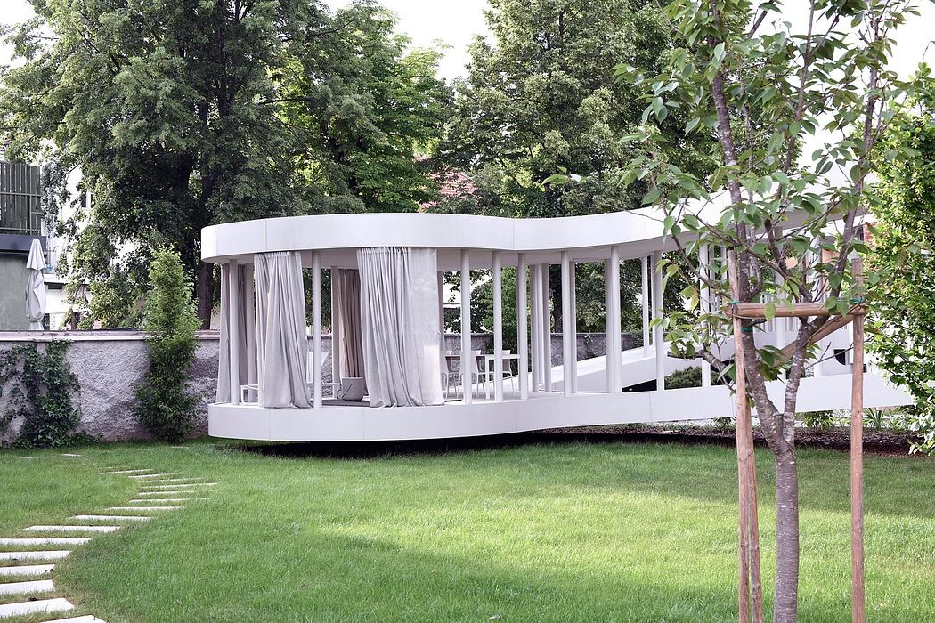 A modern, curved pavilion with white curtains stands in a lush, green garden.
