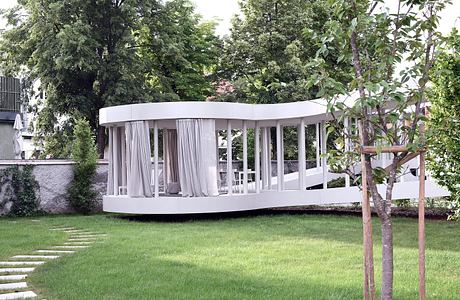 A modern, curved pavilion with white curtains stands in a lush, green garden.