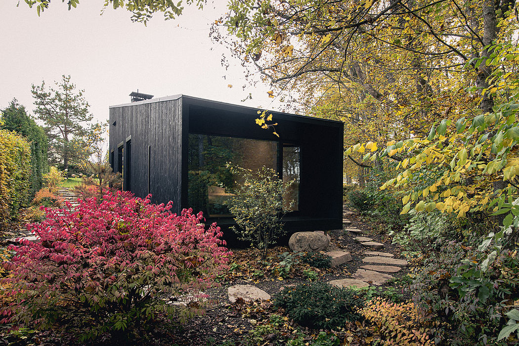 A modern black cabin nestled in a vibrant autumn garden, with glass walls and a rustic path.