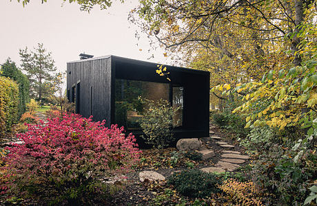 A modern black cabin nestled in a vibrant autumn garden, with glass walls and a rustic path.