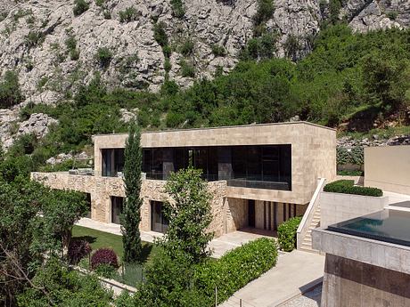 A modern, multi-story residential building with stone facade and expansive windows, set amidst lush vegetation.