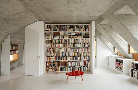 A spacious, minimalist attic with a floor-to-ceiling bookshelf, wooden furnishings, and concrete walls.